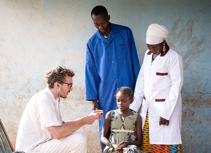 A doctor kneels to examine a young girl as two health workers in uniforms stand beside her.