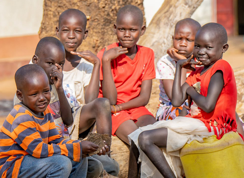 Six children sit together outdoors, several resting their chins on their hands and smiling.