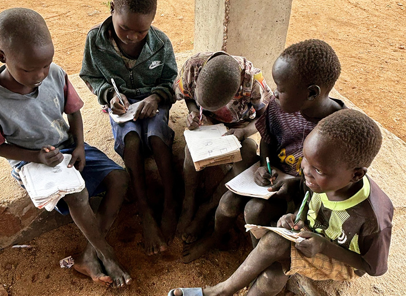 Children sit together on a ledge in the shade, writing in their notebooks.