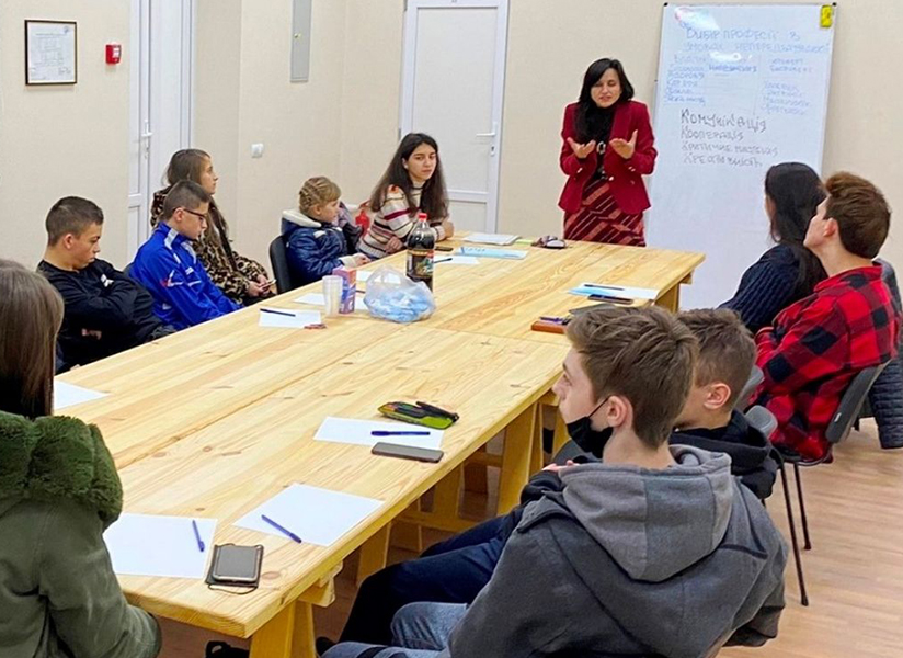 Teenagers sit around a long wooden table as a counselor leads a group session beside a whiteboard.