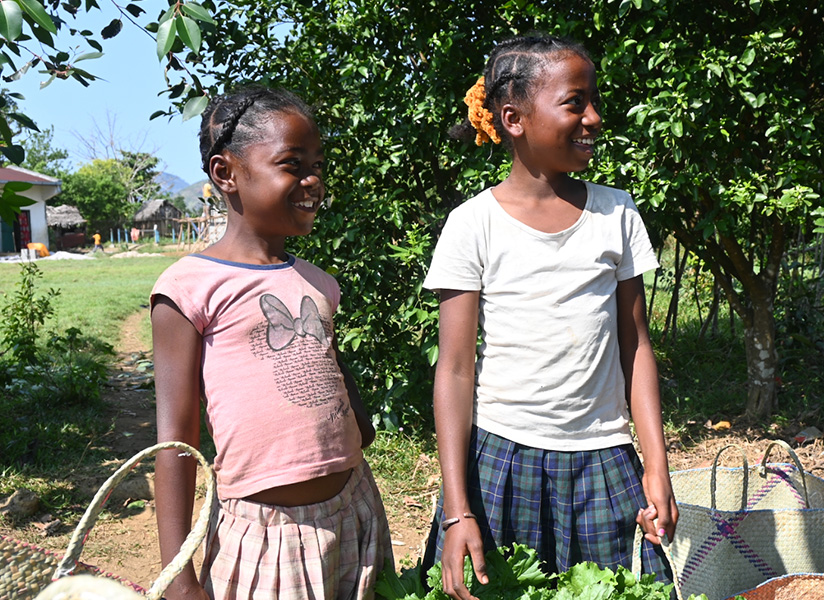 Two smiling schoolgirls stand outdoors near a garden as someone holds a hanging weighing scale.