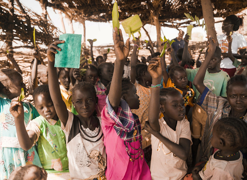 A large crowd of children raise their hands, some holding up notebooks, at an outdoor gathering.