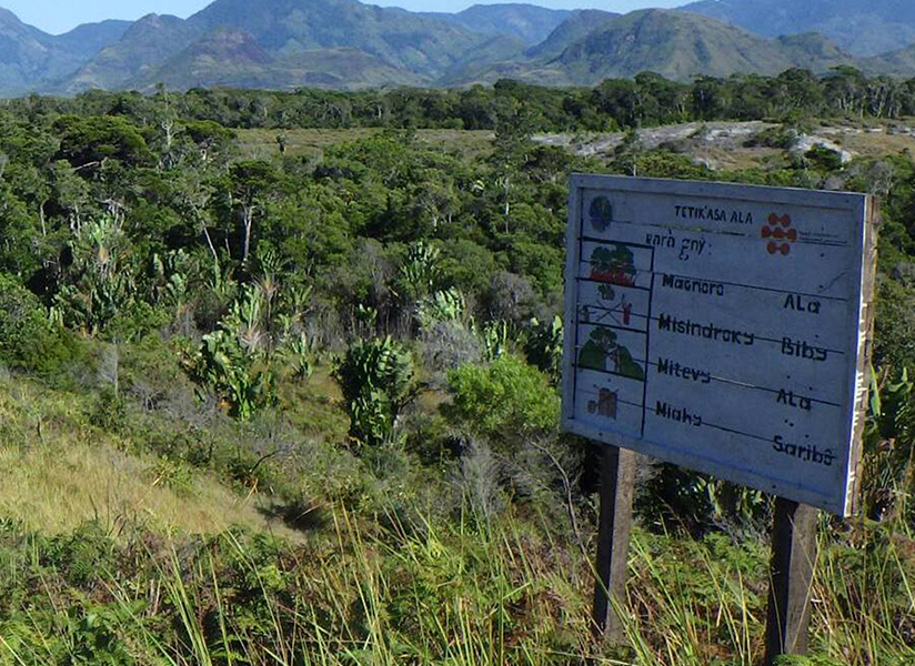 A weathered signboard stands on a green, scrubby hillside with mountains in the distance.