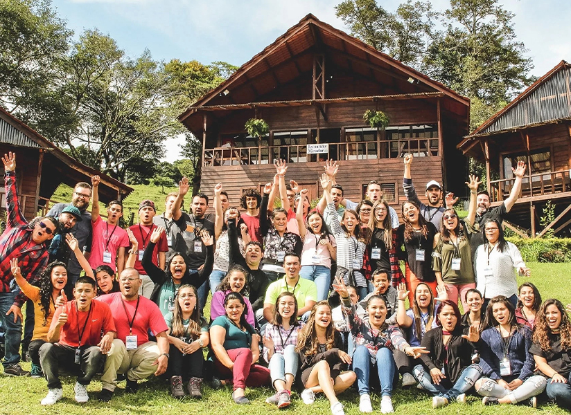 A large group of smiling community leaders pose on a lawn in front of wooden buildings, many raising their arms.