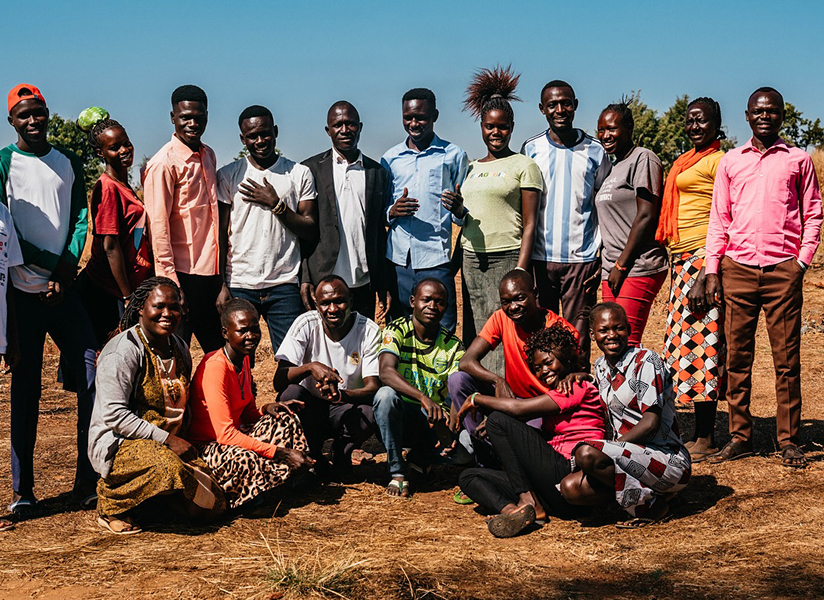 A large group of young teachers and trainees pose together outdoors on dry ground.