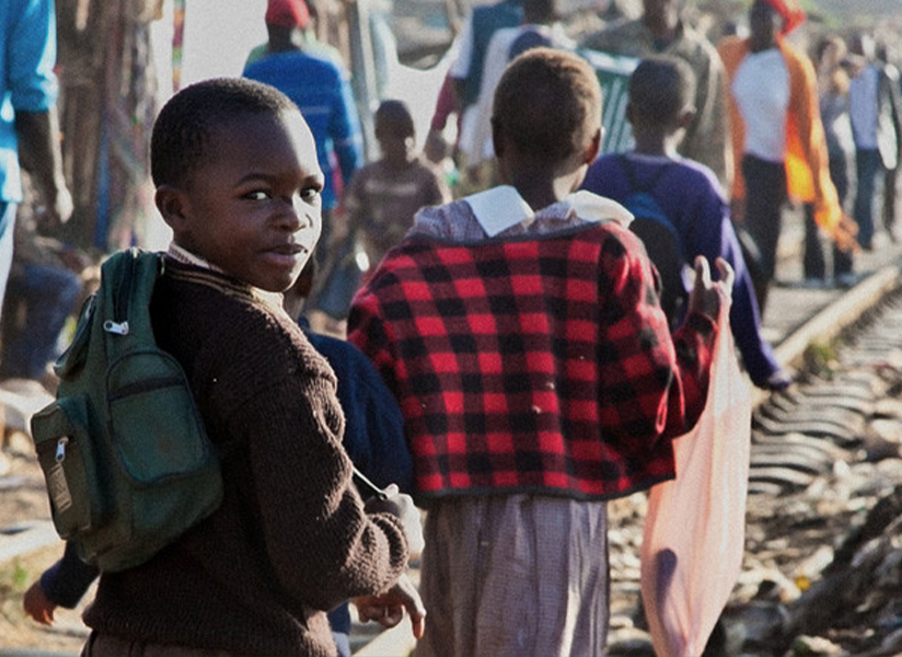 A schoolboy with a green backpack looks back over his shoulder as children walk along a railway track.