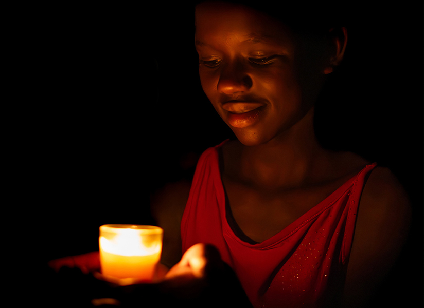 A young woman in a red top smiles softly at the candle she holds in the darkness.