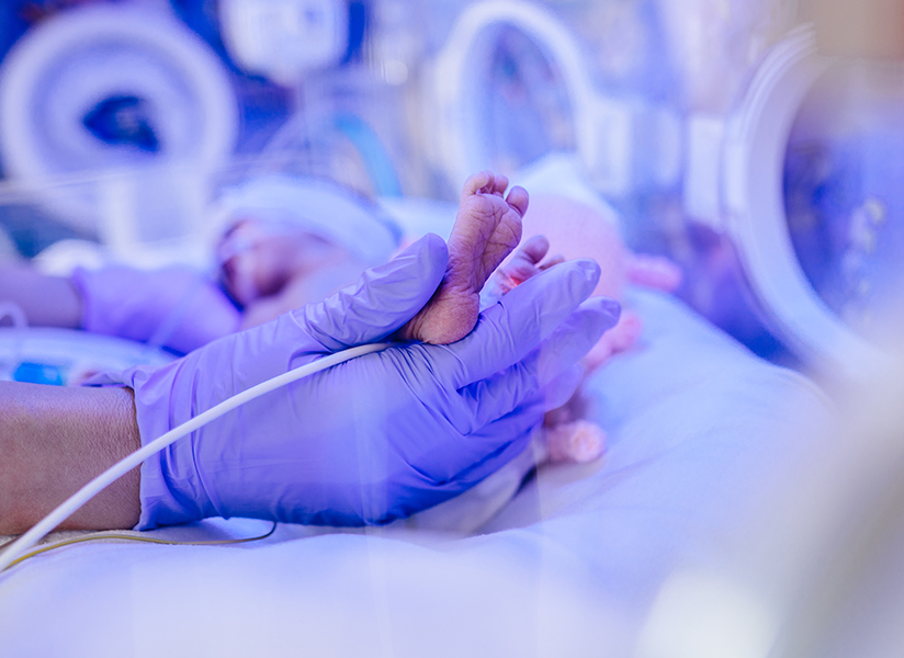 A gloved hand gently cradles a newborn's tiny foot in a neonatal incubator.