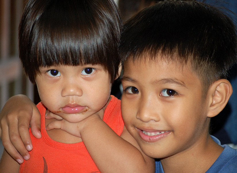 A smiling young boy wraps his arm around a toddler in an orange tank top.