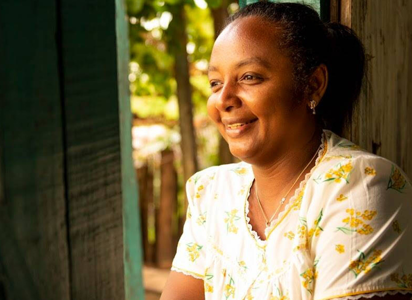 A smiling woman in a floral blouse gazes out from a sunlit doorway.