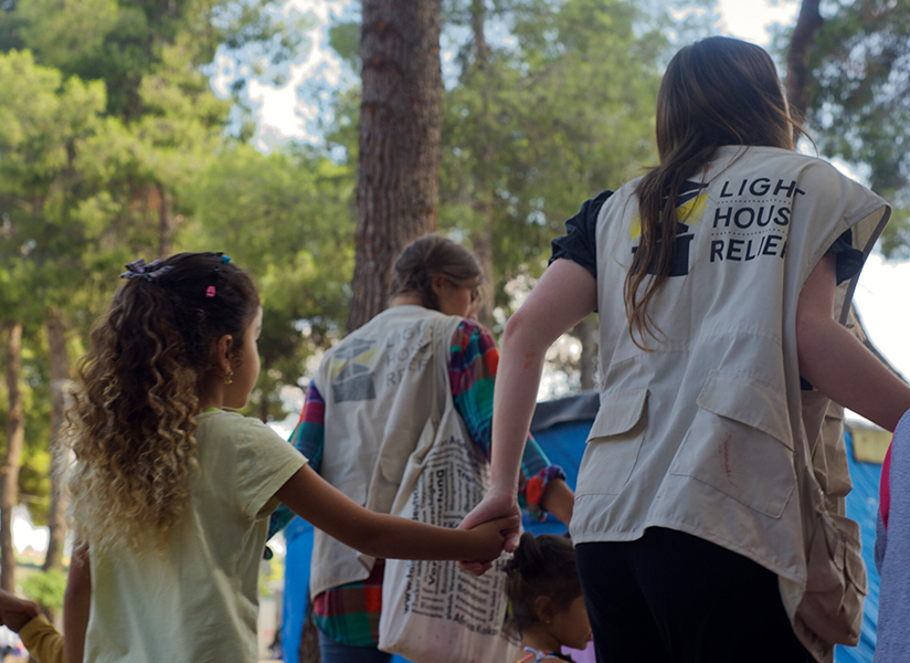 A child holds hands with Lighthouse Relief volunteers in branded vests as they walk together outdoors.