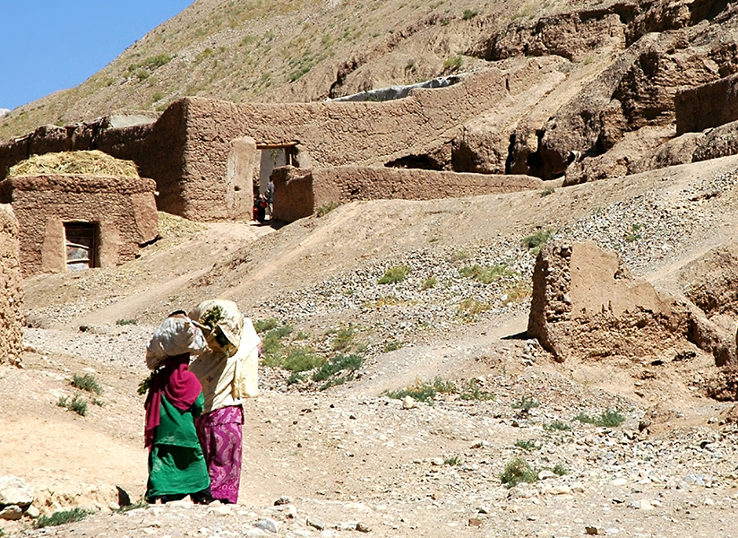 Two women in colorful clothing walk along a dusty path past mud-brick homes on a rocky hillside.