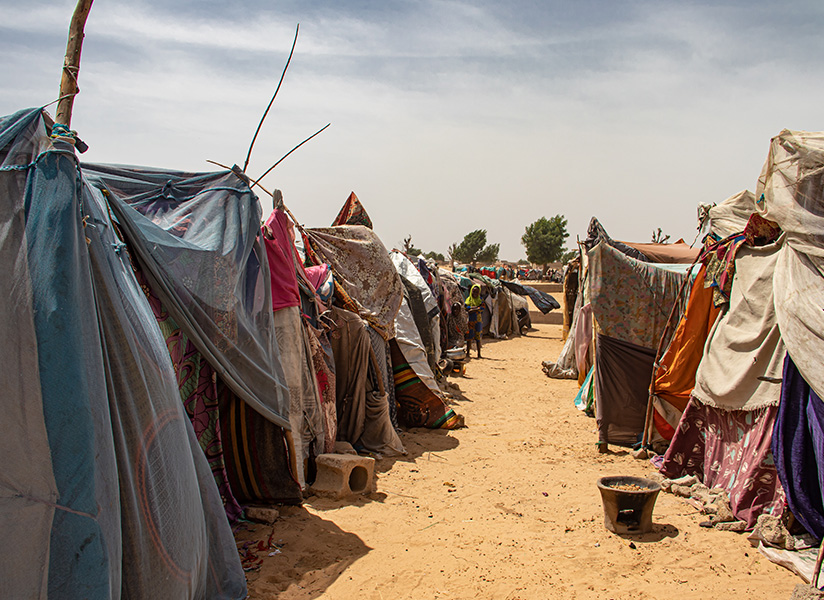 Makeshift shelters made of tarps and fabric line a sandy path in a displacement camp.