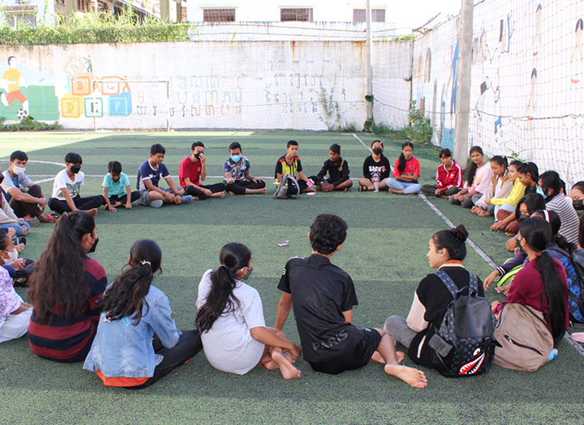 Students sit in a large circle on an outdoor football pitch for a group activity.