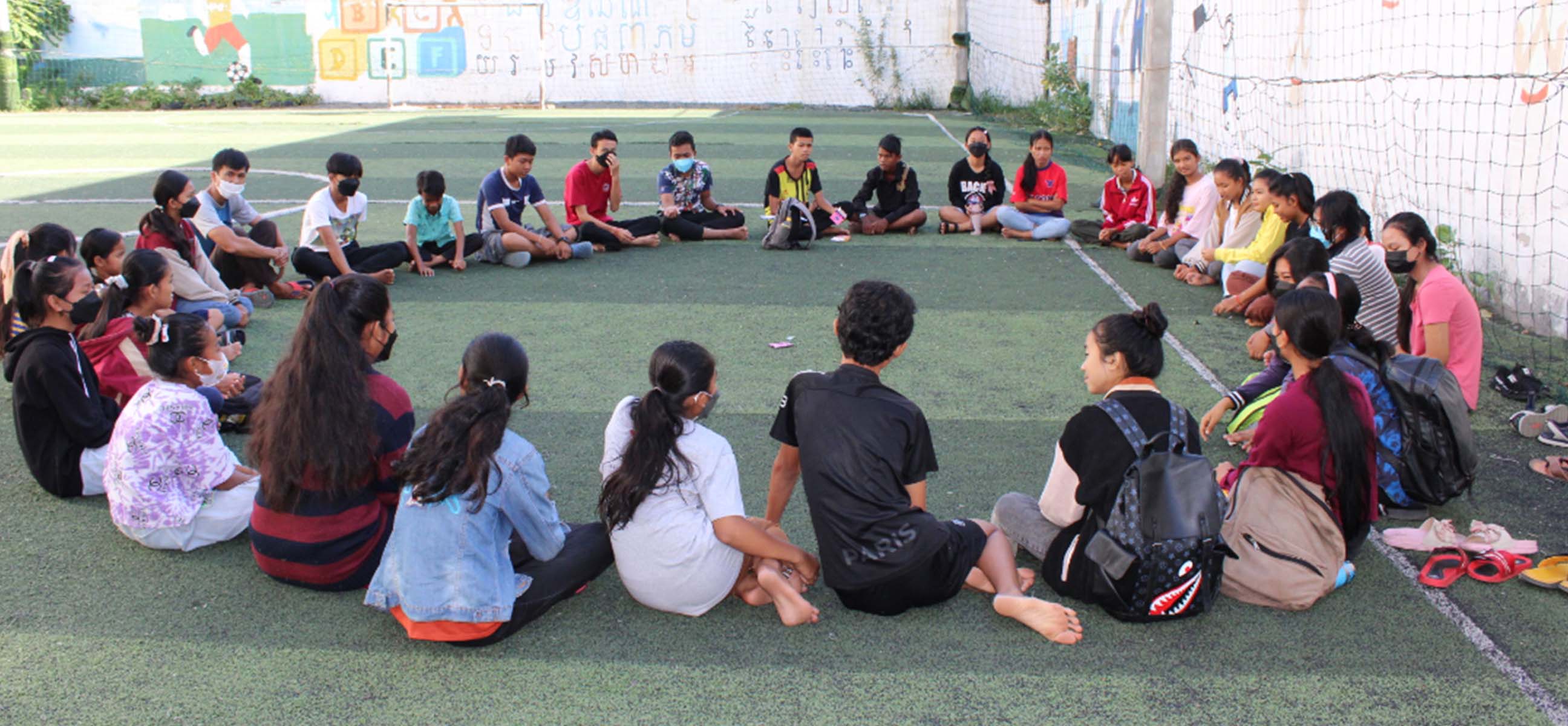Students sit in a large circle on an outdoor football pitch for a group activity.