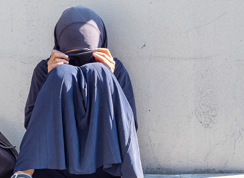 A young woman in a dark hijab sits against a wall, hiding her face behind her knees.