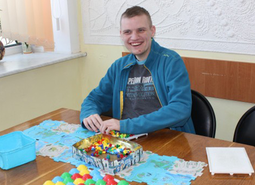 A smiling young man in a blue jacket sits at a table with a colorful craft project.
