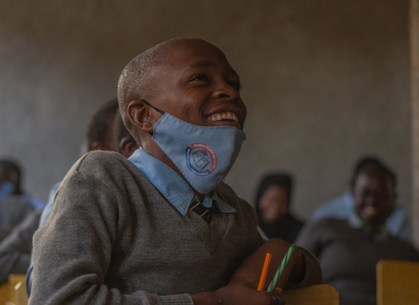 A laughing student with a face mask pulled down under his chin sits among classmates.