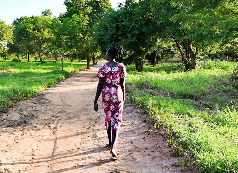 A woman in a patterned dress walks away down a dirt path between green grassy fields.