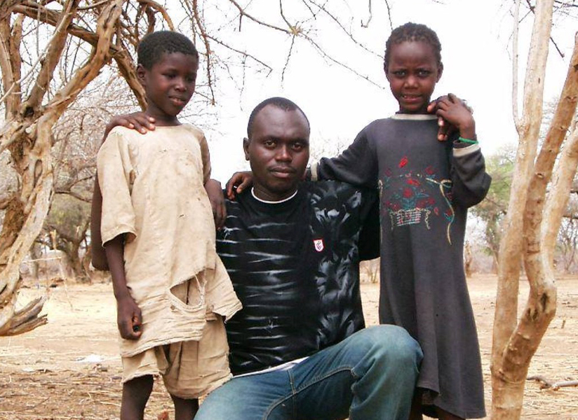 A man stands with his arms around two boys among bare trees.