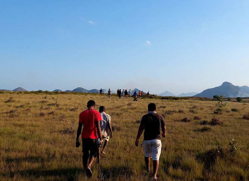 Seen from behind, three men walk across a wide, dry grassland toward distant mountains.