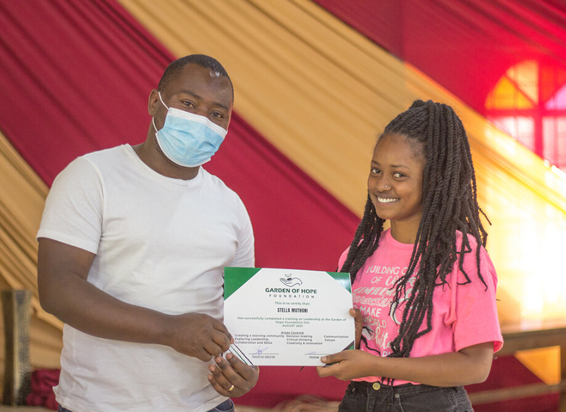 A smiling young woman receives a Garden of Hope Foundation certificate from a man in a face mask.