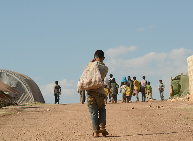 A boy carrying a large bag on his shoulder walks down a dirt road toward a group of people near tents.