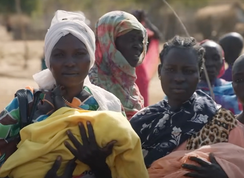 Women and young people stand together in a crowded camp, some holding bundles.