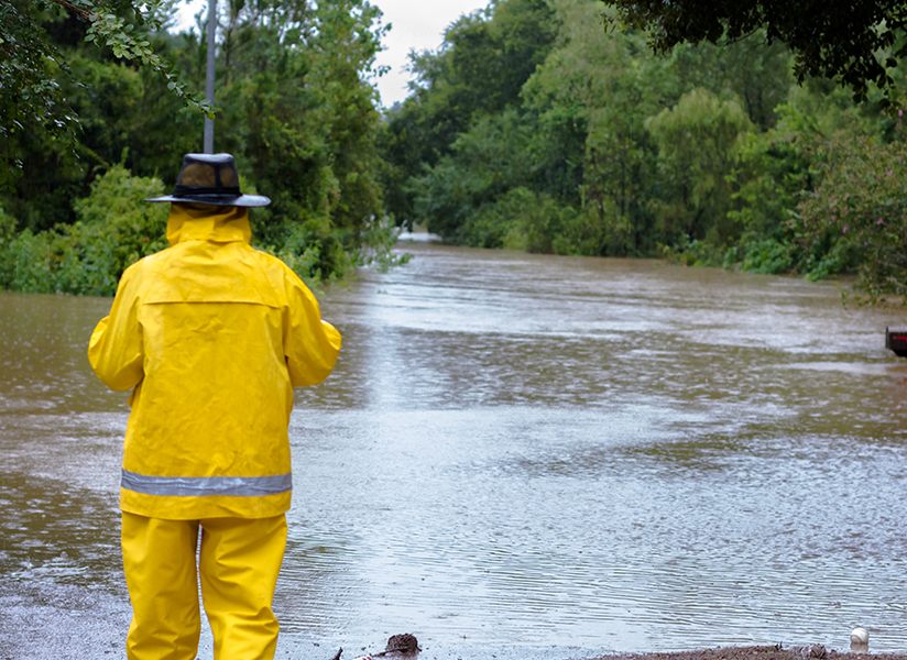 A first responder in a yellow rain suit stands looking out over a flooded road.