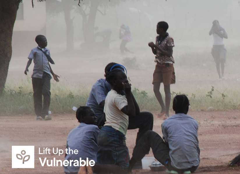 Students in school uniforms sit and stand outdoors on a hazy day, with the Lift Up the Vulnerable logo in the corner.