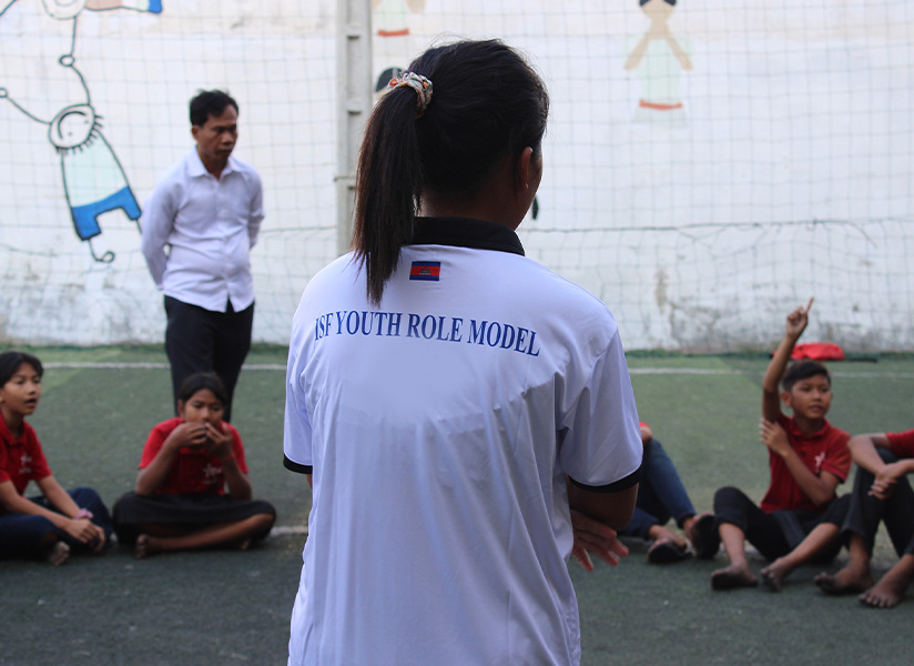 A youth leader in an "ISF Youth Role Model" shirt speaks to children seated on an outdoor pitch.