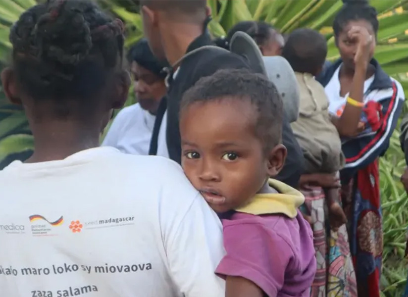 A toddler peers over the shoulder of a woman carrying him, among a crowd of mothers and children.