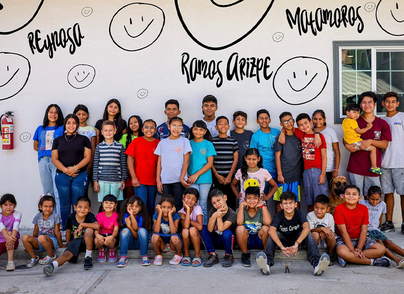 A large group of children and teenagers pose in front of a wall painted with smiley faces and the words "Ramos Arizpe."