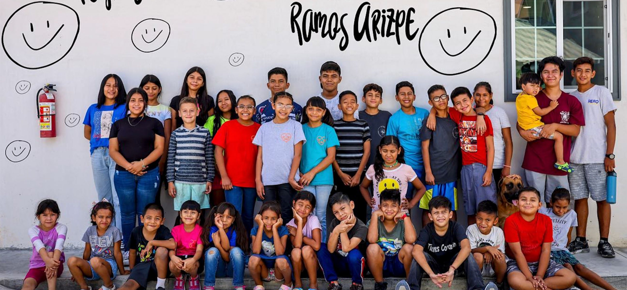 A large group of children and teenagers pose in front of a wall painted with smiley faces and the words "Ramos Arizpe."