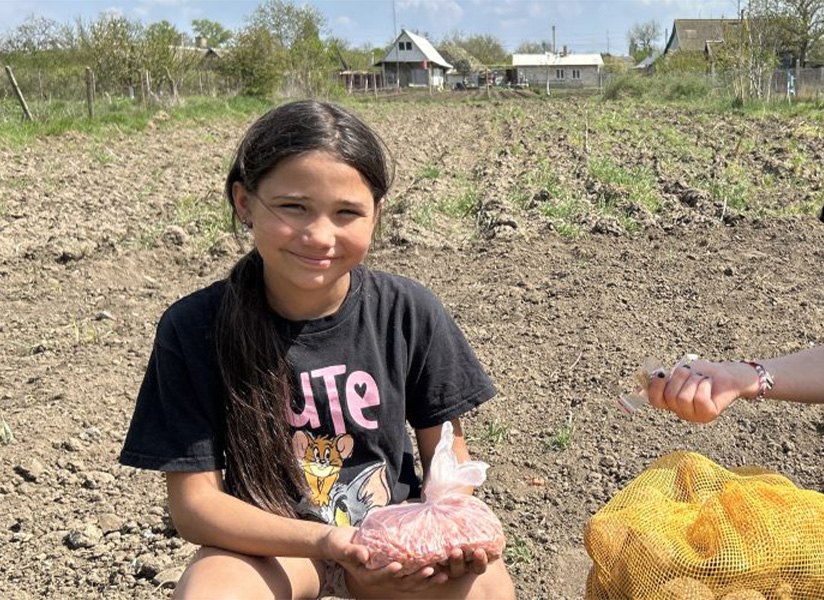 A smiling girl sits in a plowed field holding a bag of seed potatoes.