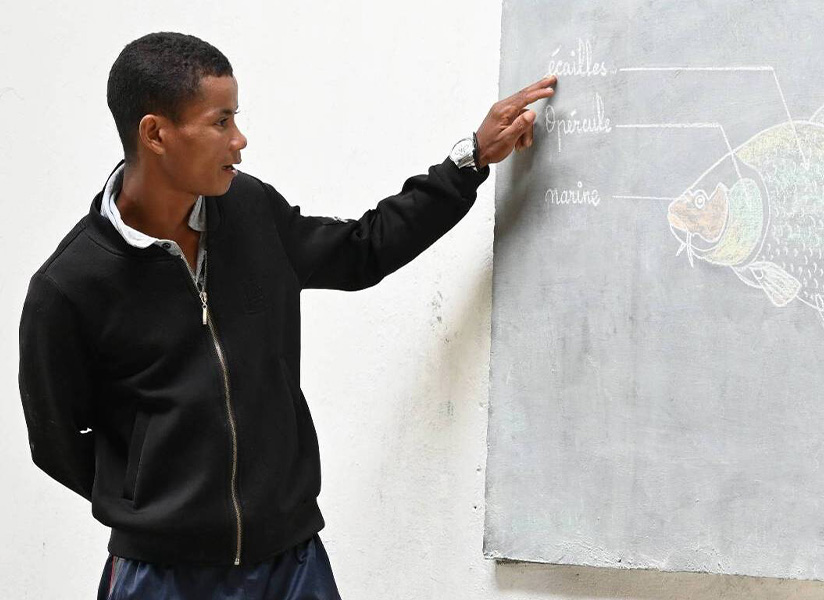 A teacher points to a chalk drawing of a fish on a classroom blackboard.