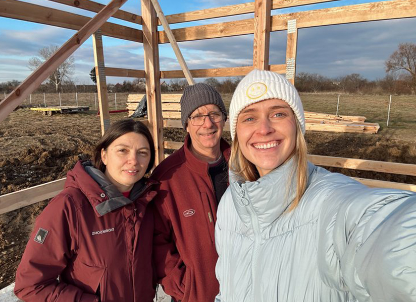 Three smiling people pose for a selfie in front of a timber frame under construction.
