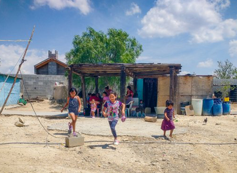 Young girls run and play in a dusty yard in front of a simple home with a shaded porch.