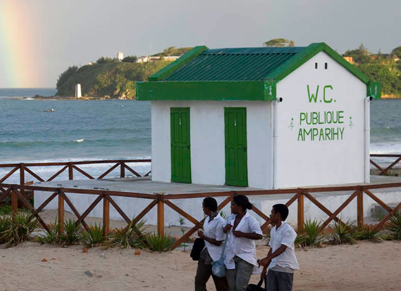 Students walk past a small public restroom building on a coastal path, with a rainbow over the sea.