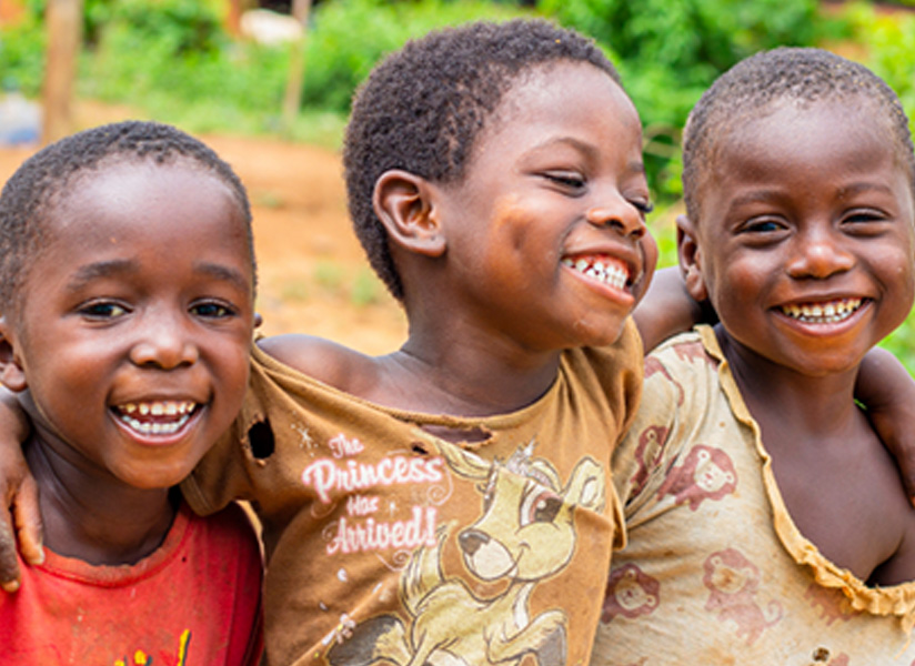 Three grinning children lean their heads together outdoors.