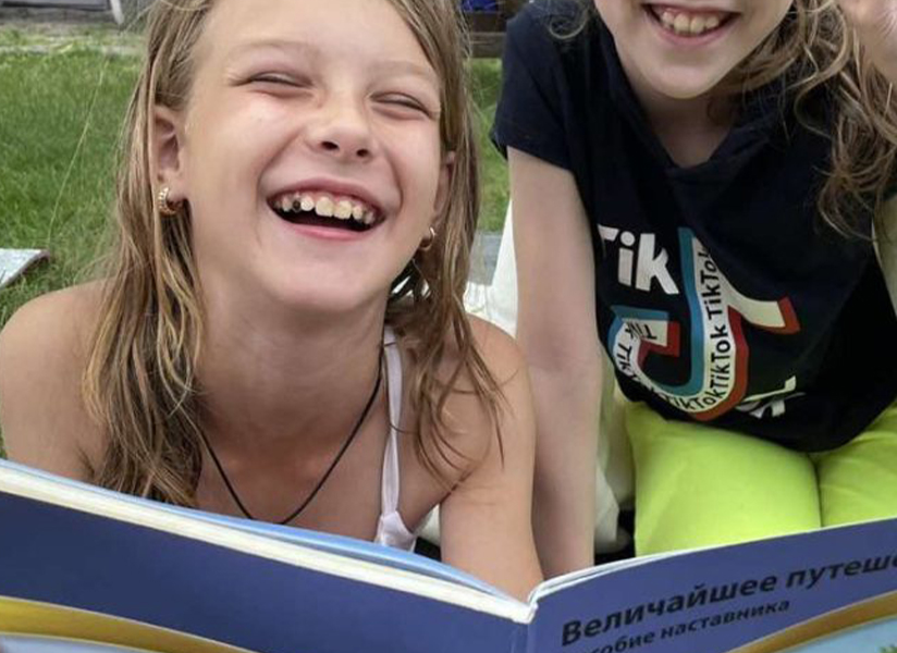Two laughing girls lie on the grass reading a picture book together.