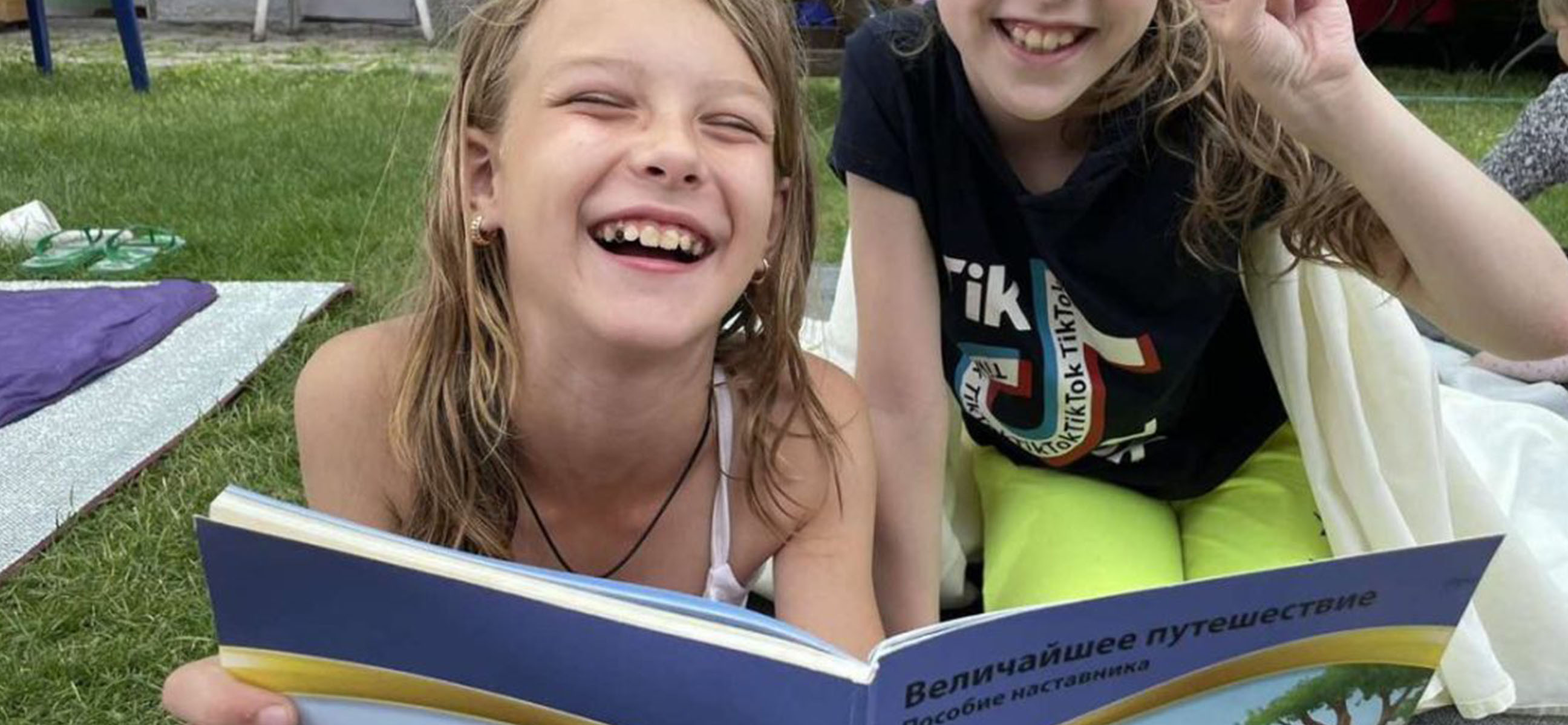 Two laughing girls lie on the grass reading a picture book together.