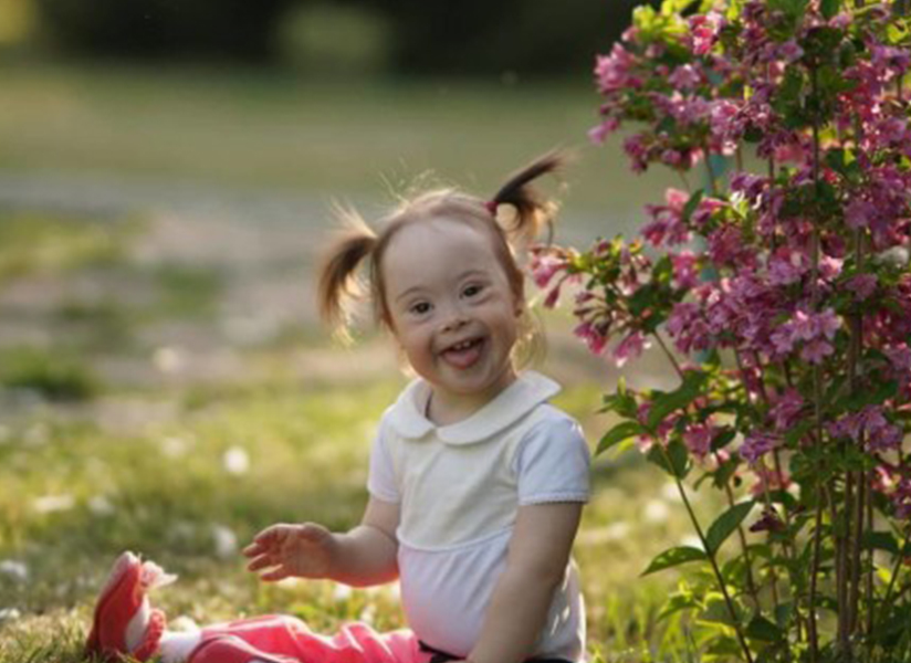 A smiling toddler with pigtails sits on the grass beside a flowering bush.