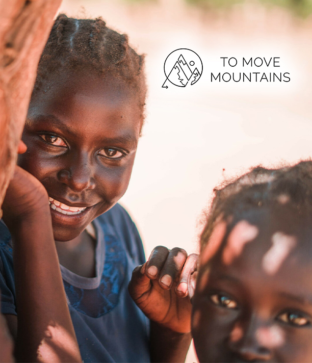 Two smiling young children peek around a doorway, beside the To Move Mountains logo.