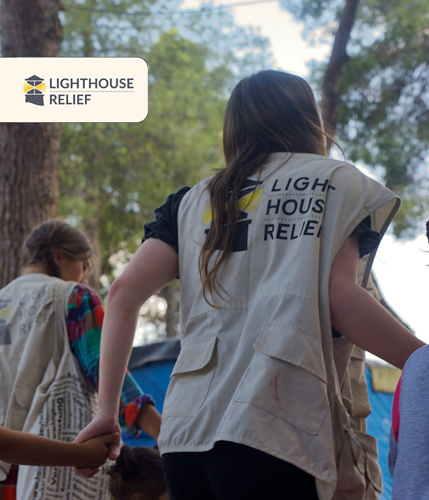 A child holds hands with Lighthouse Relief volunteers in branded vests as they walk together outdoors, with the Lighthouse Relief logo.