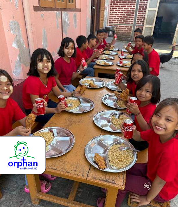 Children in red shirts smile as they share a meal together at a long outdoor table, beside the Orphan Relief Effort logo.
