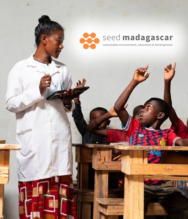 A teacher in a white coat stands among wooden desks as her students raise their hands, beside the SEED Madagascar logo.