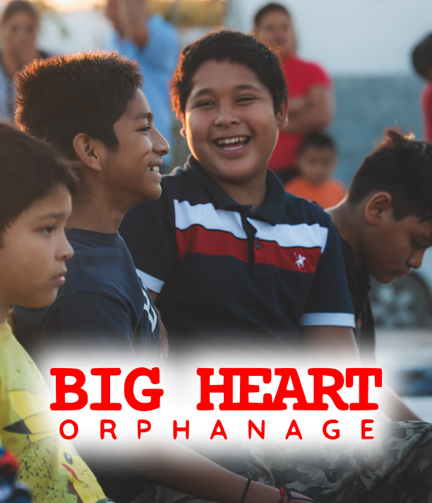 Smiling boys sit together outdoors in the late-afternoon sun, above the Big Heart Orphanage logo.