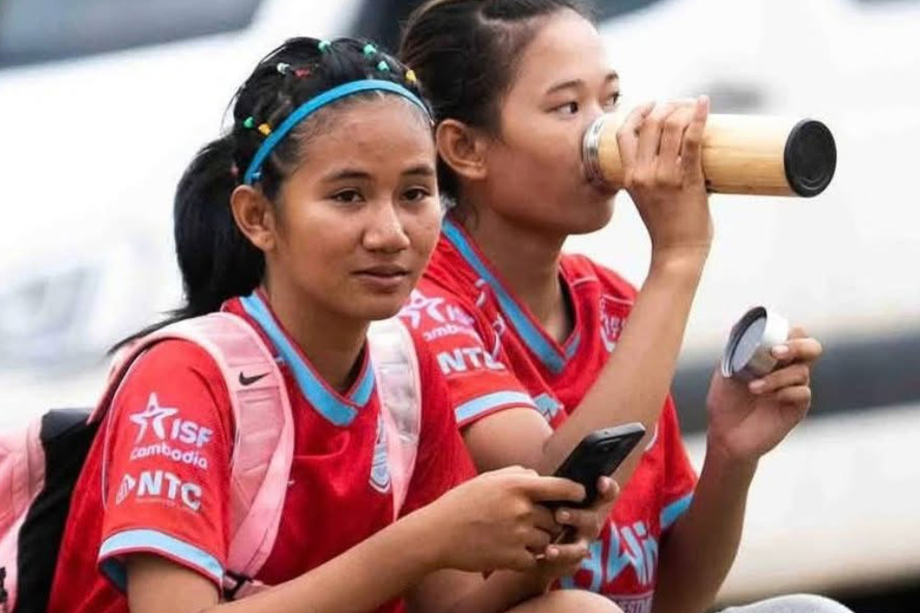 Two young footballers in red ISF Cambodia kits sit side by side, one drinking from a bamboo flask and the other holding a phone.