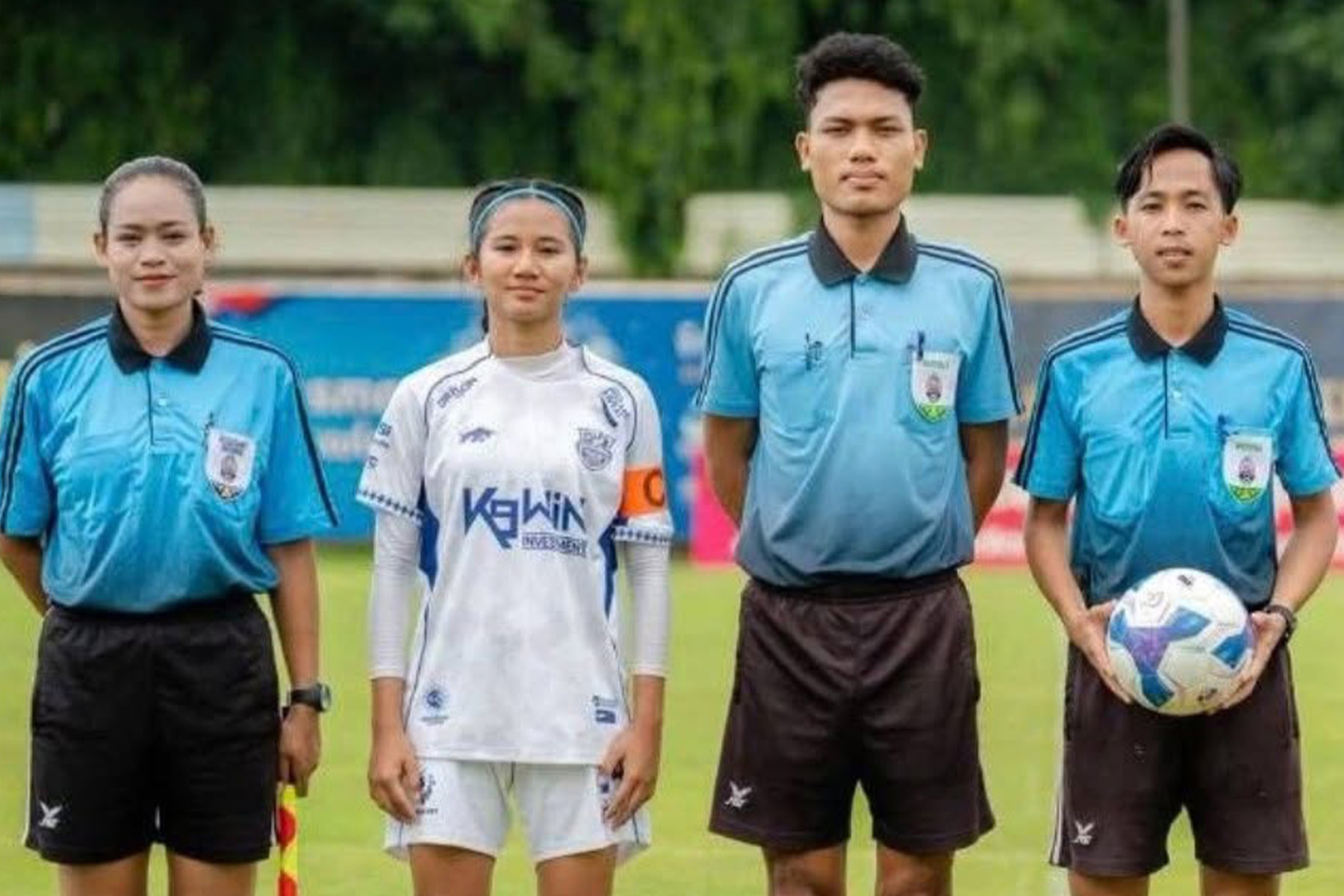 A young footballer in a white and blue kit stands with three match officials in pale blue shirts before kickoff, one holding the match ball.
